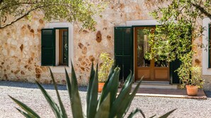 Un edificio rústico de piedra con dos ventanas, cada una con contraventanas verdes, y una puerta de madera. Hay plantas en macetas frente a la puerta.