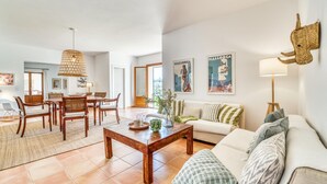 Amplia sala de estar con una mesa de comedor de madera, un sofá y una mesa de centro. Hay carteles en la pared y una lámpara a la derecha.