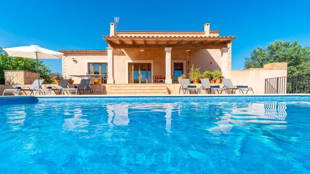 A poolside area with a clear blue pool, lounge chairs, and a building with a tiled roof and columns. - Awesome home in Alqueria Blanca (Alqueria Blanca)