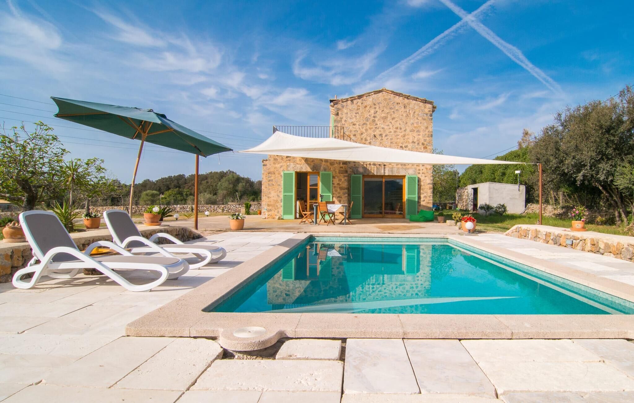 A pool area with lounge chairs, a stone building with green shutters, and a clear blue sky.