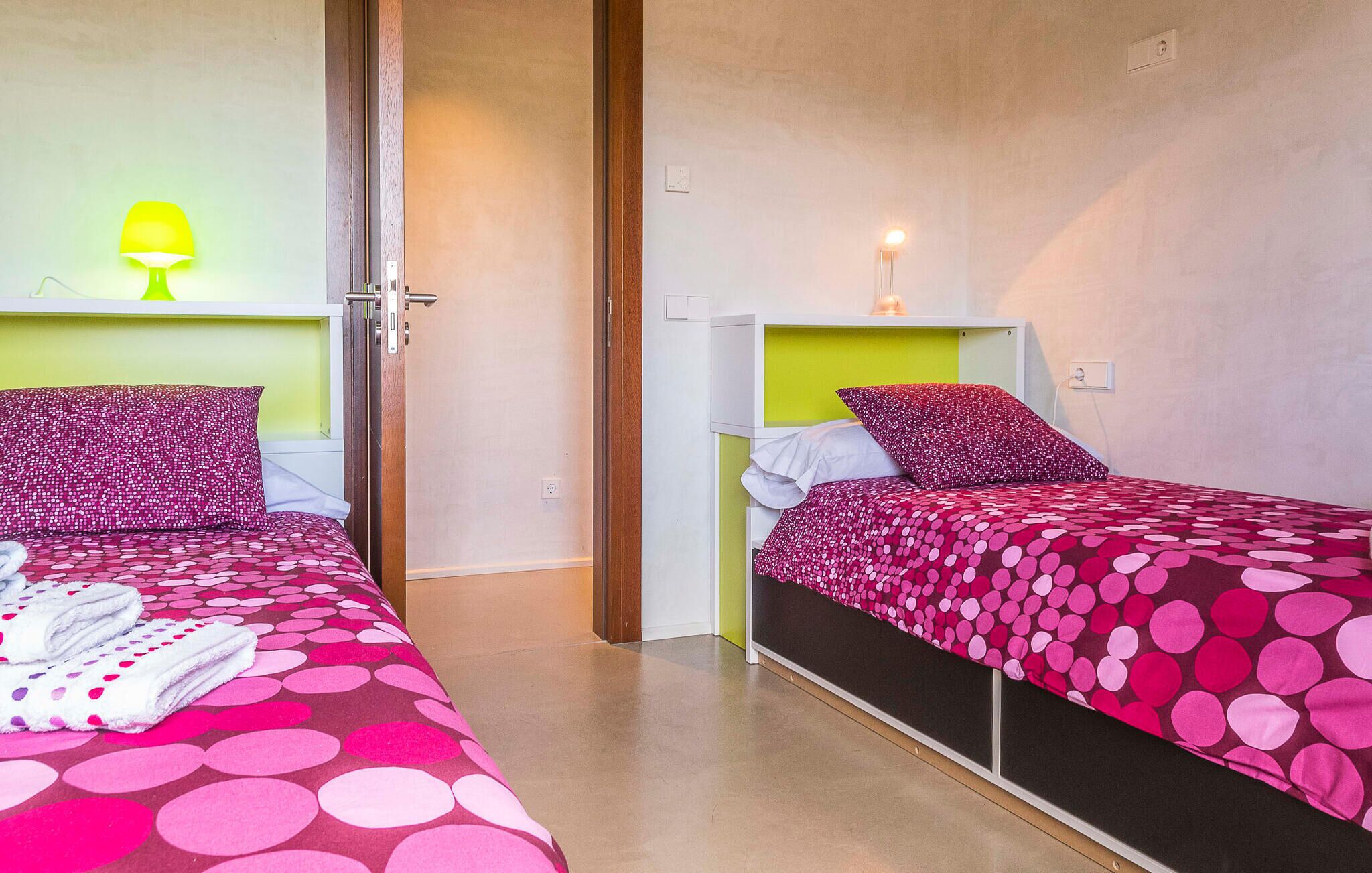 Ein Hotelzimmer mit zwei Betten, jedes mit einer dezenten, gepunkteten Bettdecke und passenden Kissen. Links in der Ecke steht eine Lampe, rechts am Bett eine Nachttischlampe.