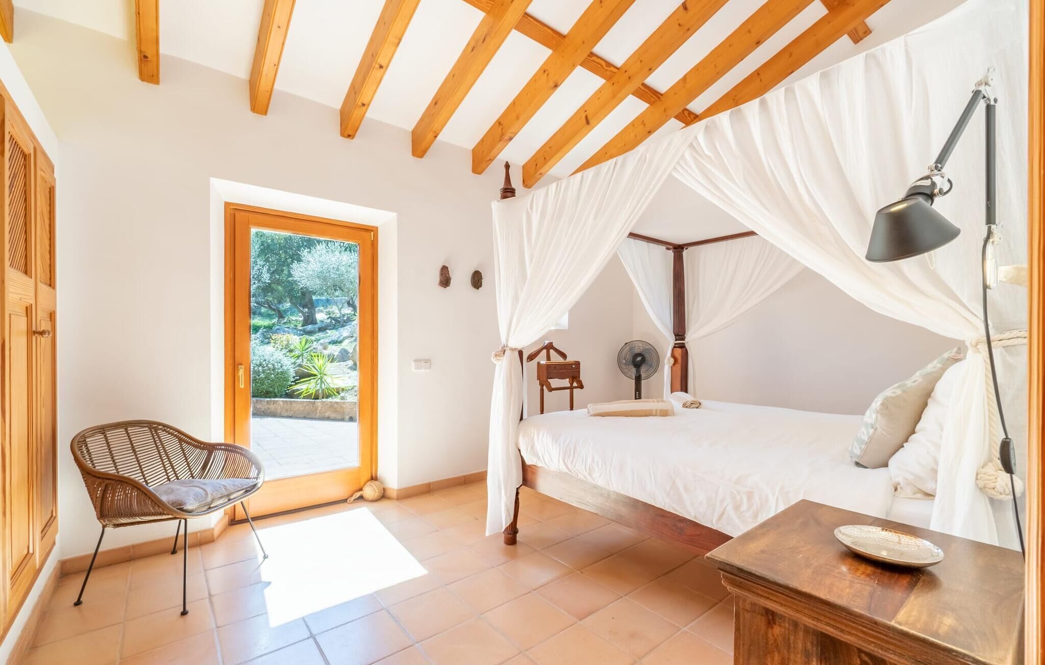 A bedroom with a four-poster bed, a bedside table, a chair, and a view of a garden through a glass door.