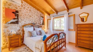 A bedroom with a stone wall, wooden ceiling, a bed with white bedding, a nightstand, a dresser, and a window with a view of palm trees. - Pet friendly home in Can Picafort (Can Picafort)
