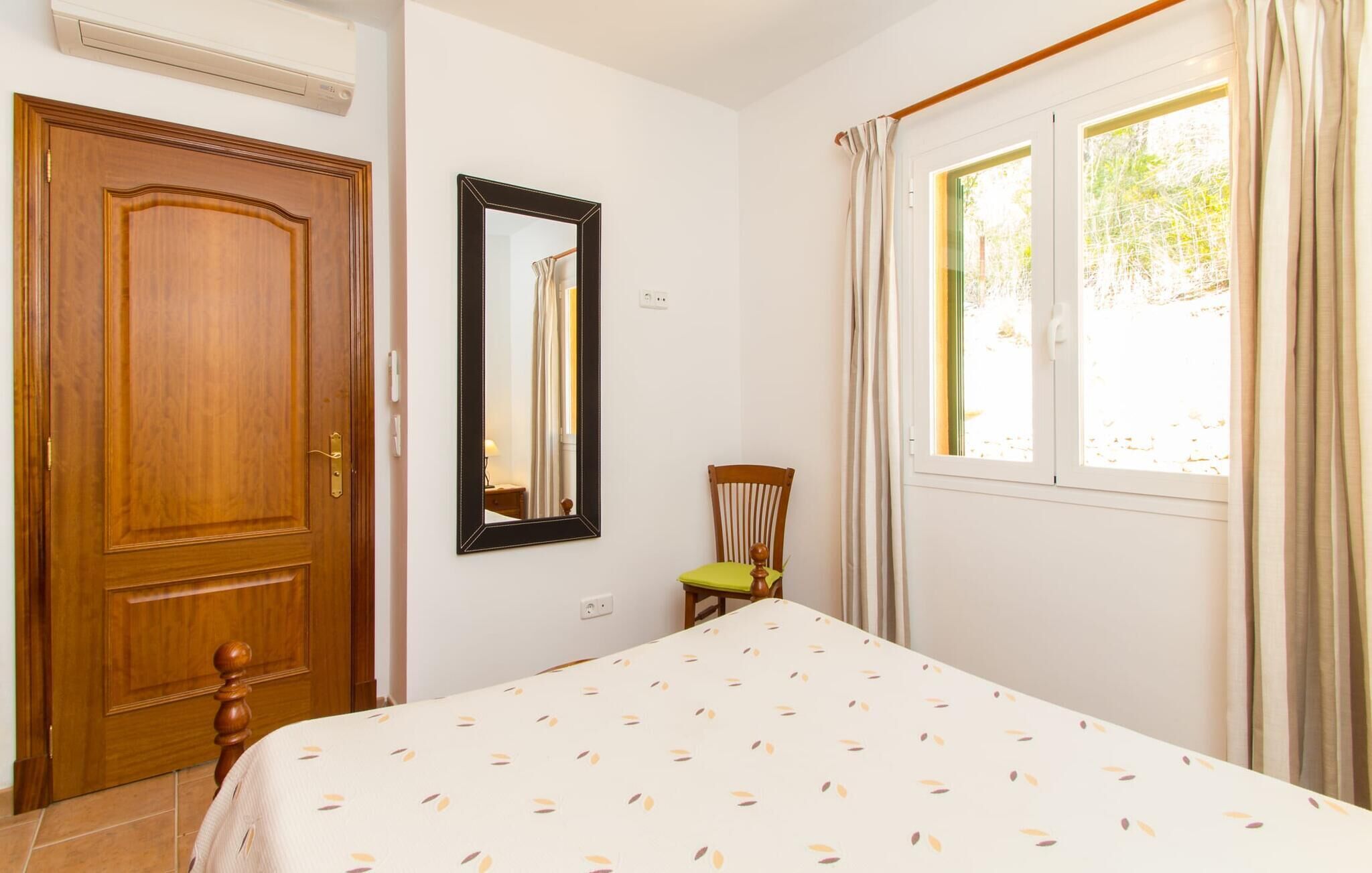 A bedroom with a wooden door, a bed with a leaf-patterned bedspread, a mirror, a chair, and a window with curtains.