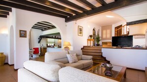 Una sala de estar con un sofá, una mesa de centro y un televisor. Se ve una escalera que lleva a una zona de cocina.