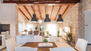 Un vaste espace de salle à manger avec une table en bois, des chaises blanches et des luminaires suspendus. On aperçoit, au fond, une cuisine équipée d’appareils modernes.