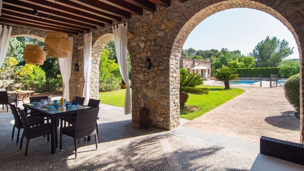 Eine Außenpatio mit steinernen Bögen, Korbmöbeln und Blick auf einen Pool und viel Grün.