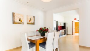 Een eethoek met een houten tafel en witte stoelen, een potplant met rode bloemen en twee ingelijste kunstwerken aan de muur.