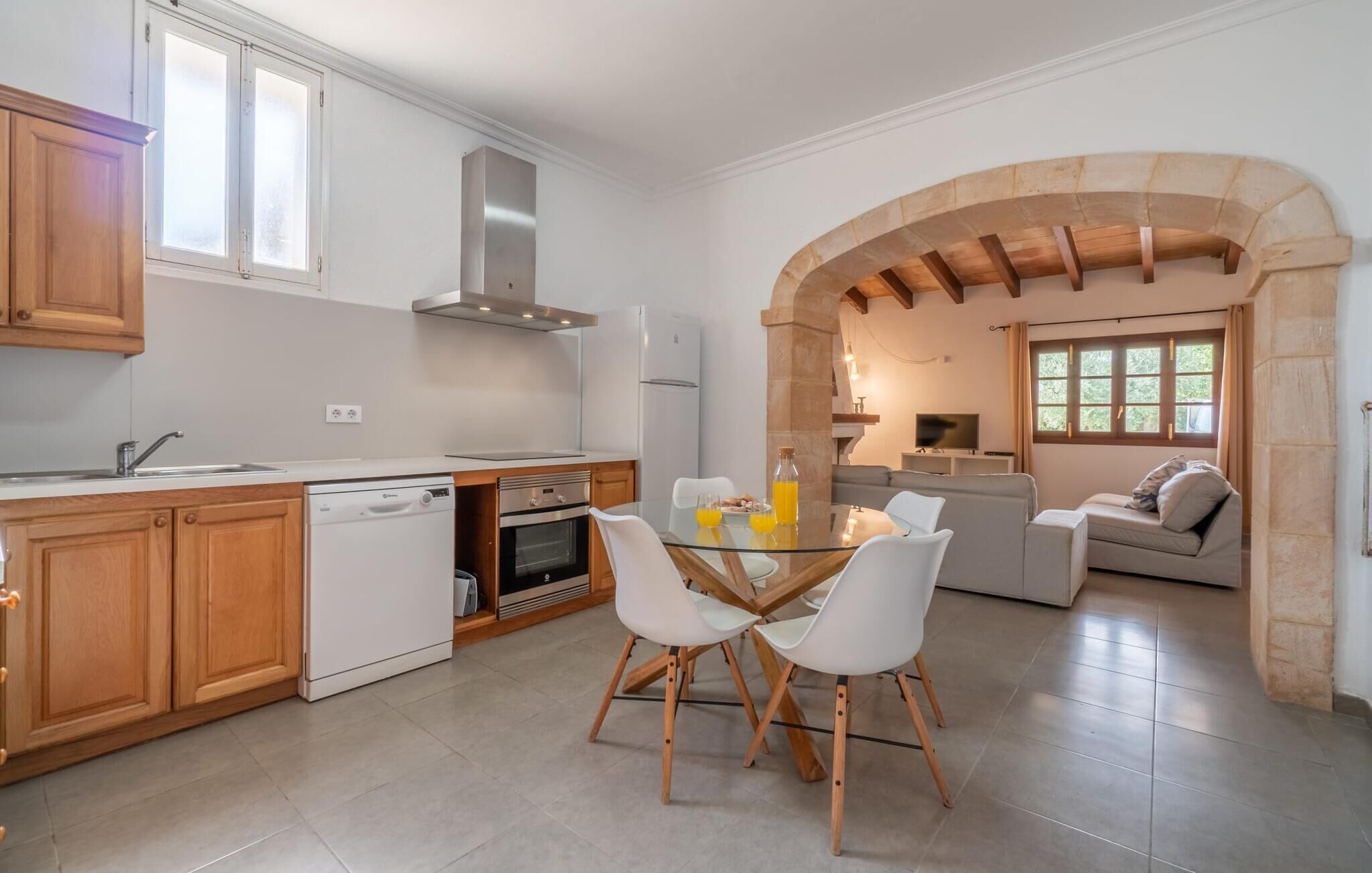 Una cocina moderna con armarios de madera, lavavajillas, horno y cocina empotrada. Hay una zona de comedor con una mesa de vidrio y sillas blancas. Un arco de entrada conduce a una sala de estar con un sofá y televisor.