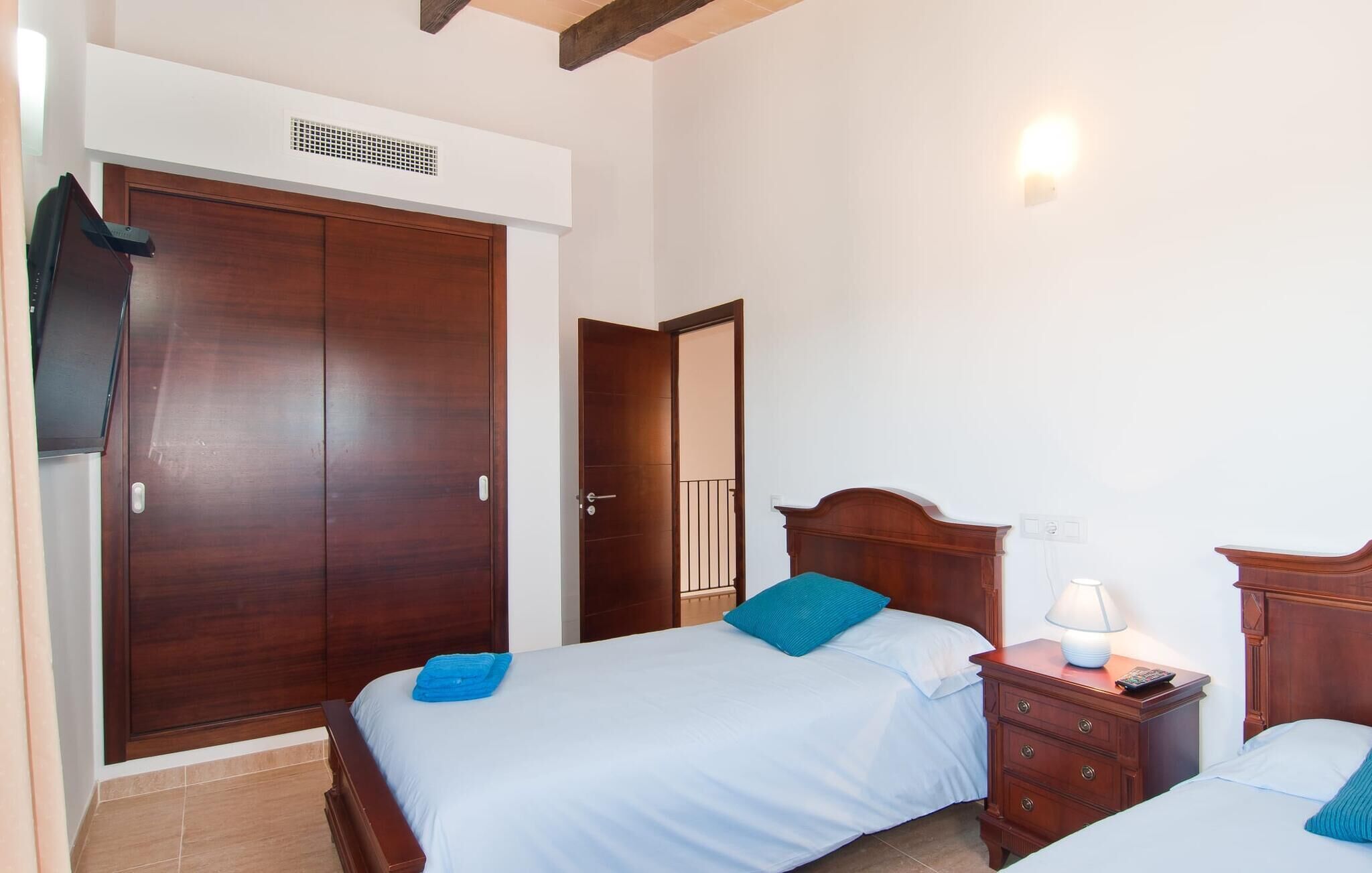 A bedroom with a large wardrobe, a bed with blue bedding, a bedside table with a lamp, and a small bedside table with a remote control.