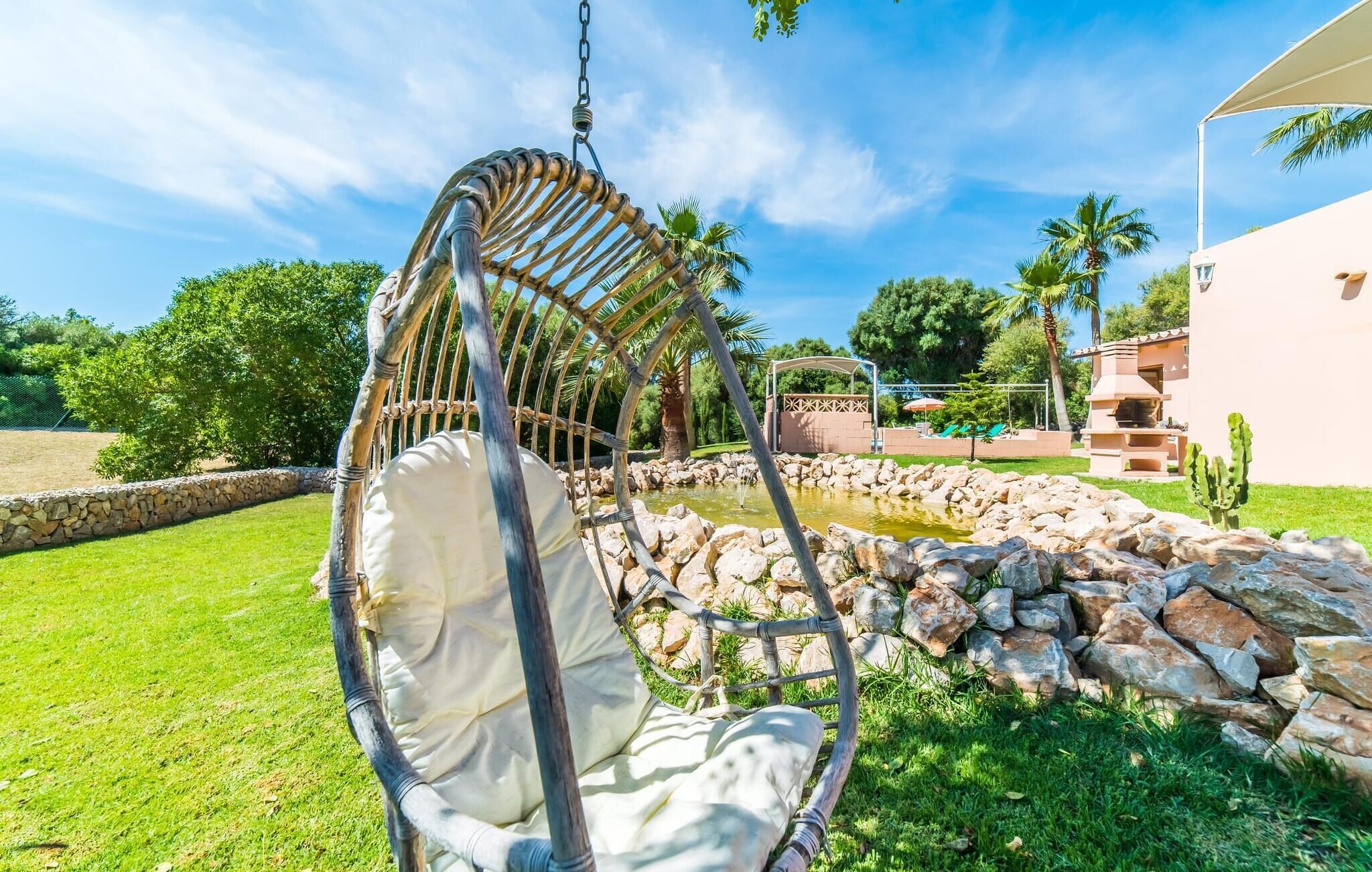 Uma cadeira suspensa em um jardim com piscina, muro de pedra e um edifício ao fundo.