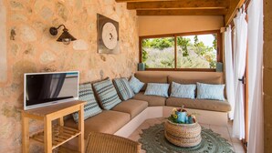 A cozy living area with a stone wall, wooden ceiling beams, a large window with a view of greenery, a television on a wooden stand, and a woven ottoman. - Beautiful home in Petra with private swimming pool, can be inside or outside (Petra)