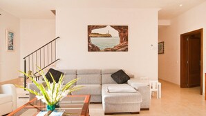 Ein Wohnzimmer mit grauem Sofa, einem Couchtisch mit einer Vase weißer Blumen, einem Gemälde einer Küstenlandschaft und einer Treppe.