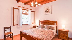 Ein Schlafzimmer mit einem hölzernen Bett, einem Nachttisch, einem Sessel, einem Fenster mit Vorhängen und einer Deckenleuchte.