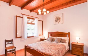 Ein Schlafzimmer mit einem hölzernen Bett, einem Nachttisch, einem Sessel, einem Fenster mit Vorhängen und einer Deckenleuchte.