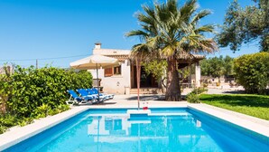 Ein Swimmingpool mit blauen Fliesen, umgeben von einer weißen Poolterrasse. Blaue Liegestühle und ein weißer Sonnenschirm sind vorhanden. Eine Palme und weitere Grünpflanzen sind zu sehen.