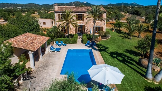 Una amplia casa de piedra con piscina, rodeada de exuberante vegetación y palmeras.