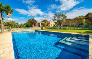 Une piscine rectangulaire à l’eau d’un bleu cristallin, entourée d’une clôture en bois peinte en blanc, d’un bâtiment en pierre et d’une végétation luxuriante.