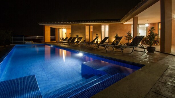 En swimmingpool med liggestole og krukkeplanter, omgivet af en bygning med store vinduer og varm belysning indeni.