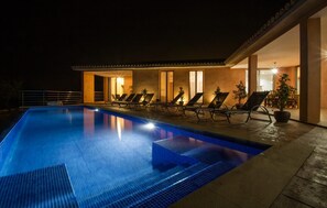 En swimmingpool med liggestole og krukkeplanter, omgivet af en bygning med store vinduer og varm belysning indeni.