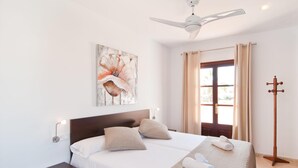 Una cama doble con dos almohadas, un ventilador de techo, un cuadro, una ventana con cortinas y una lámpara de pie de madera.