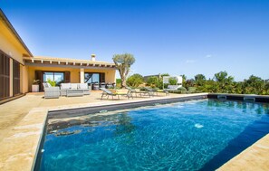 Une piscine entourée de chaises longues et d’un espace détente avec canapés, le tout baigné de verdure et surplombé par un ciel dégagé.