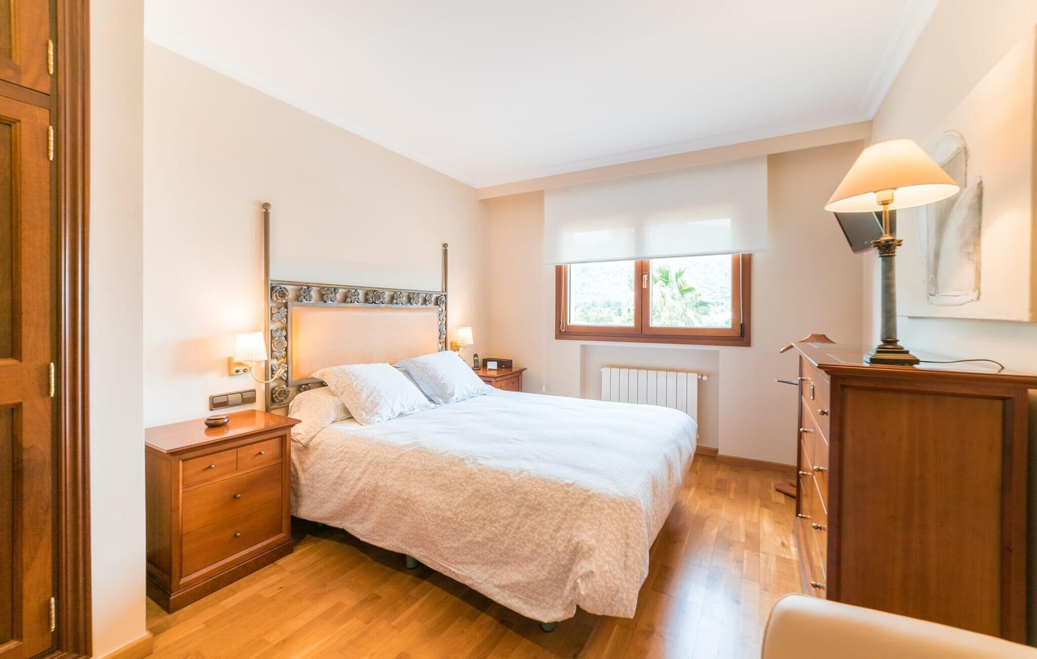 A bedroom with a bed, wooden nightstands, a dresser, a window with a view, and a lamp.