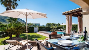 An outdoor dining area with a table set for two, a large umbrella, and a view of a swimming pool and mountains in the background. - Nice home in Alcudia with private swimming pool, can be inside or outside (Alcudia)