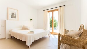 Een slaapkamer met een bed, een rieten stoel, een raam met gordijnen en een kleine plant op het nachtkastje.