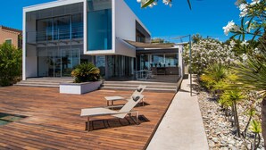 Une maison moderne à deux étages, dotée d’une terrasse en bois, d’un espace salon extérieur et d’un jardin agrémenté de fleurs blanches.