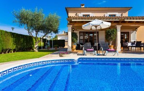 Un espace piscine avec des chaises longues et des parasols, une terrasse couverte et une pelouse bien entretenue.