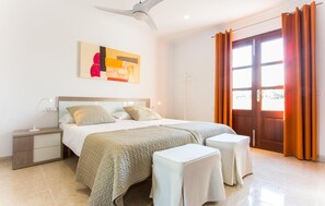 Une chambre à coucher avec un lit, des tables de chevet, un ventilateur de plafond, un tableau et une vue par une fenêtre avec des rideaux.