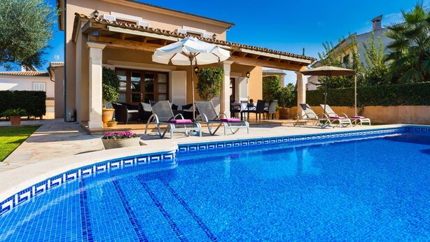 En swimmingpool med liggestole og parasoller, en overdækket terrasse og en velplejet have.