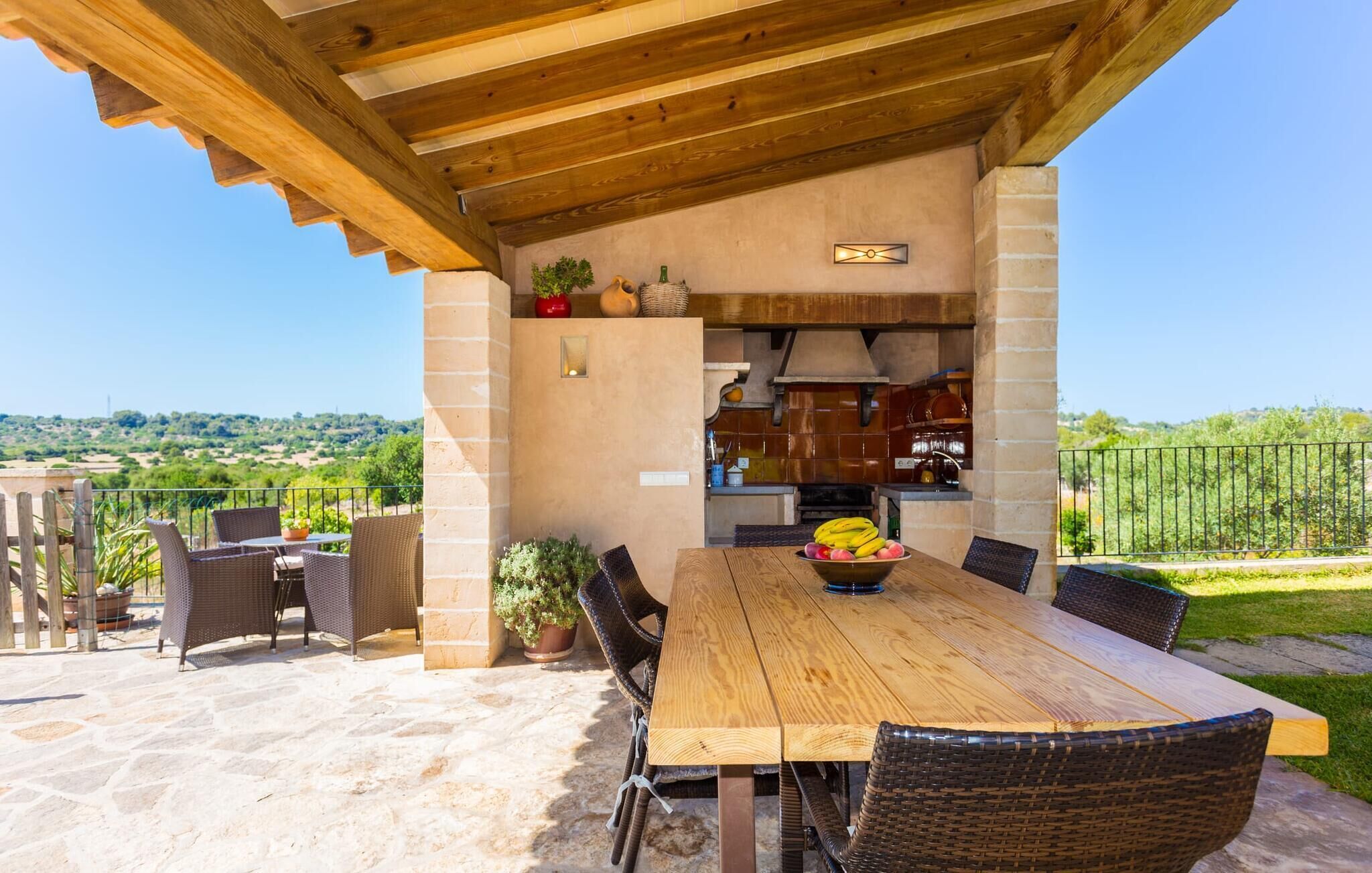 Eine Terrassensitzgruppe im Freien mit einem Holztisch, Korbstühlen, Steinboden und Blick ins Grüne.