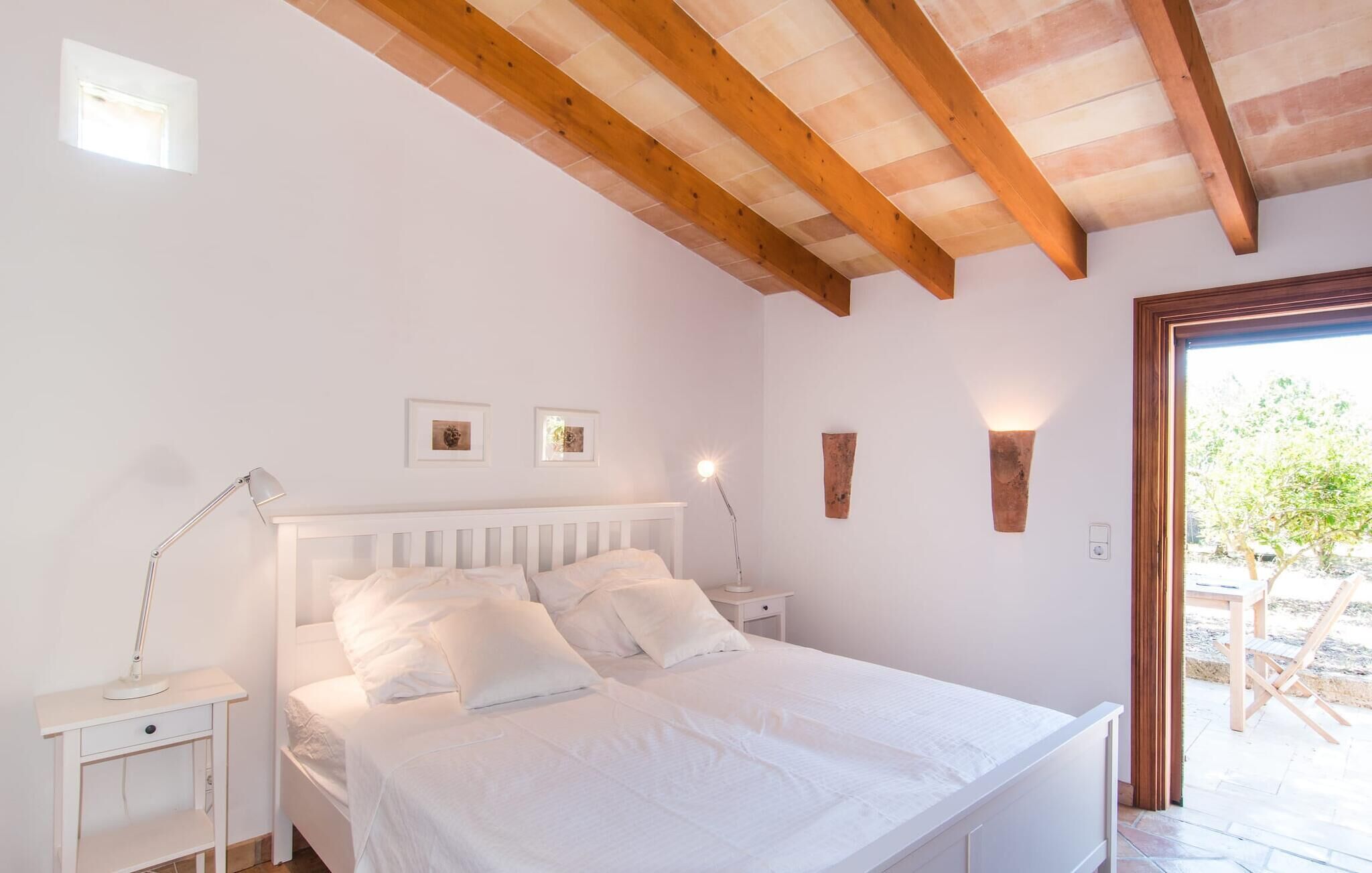 A bedroom with a wooden ceiling, a white bed, bedside lamps, and a door leading to an outdoor area.
