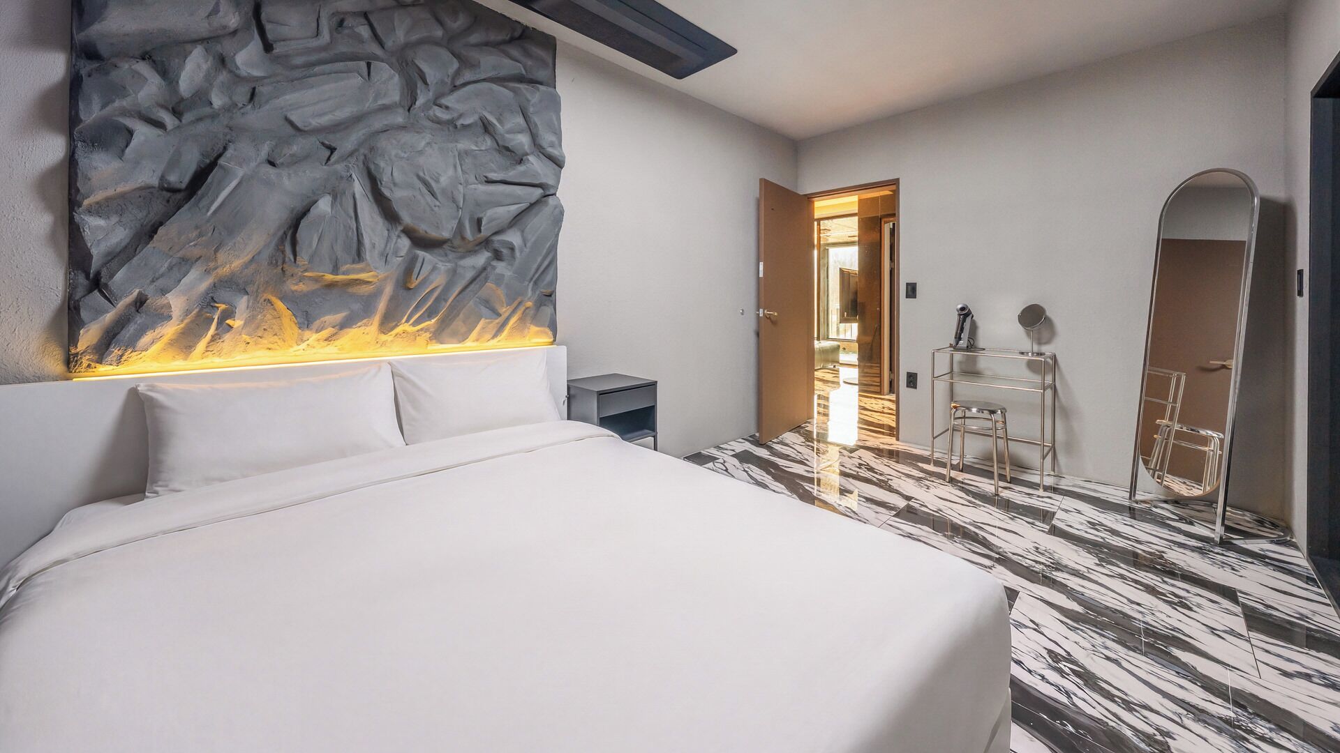 Una habitación de hotel moderna con una cama grande, una mesita de noche, un escritorio con una silla, un espejo y un elemento decorativo en la pared con textura.