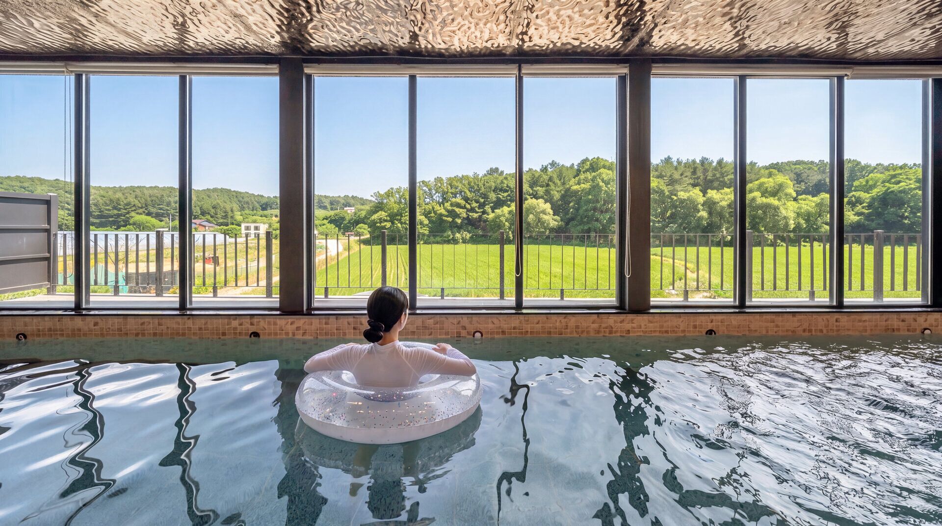 A person in a pool with a view of a green landscape through large windows.