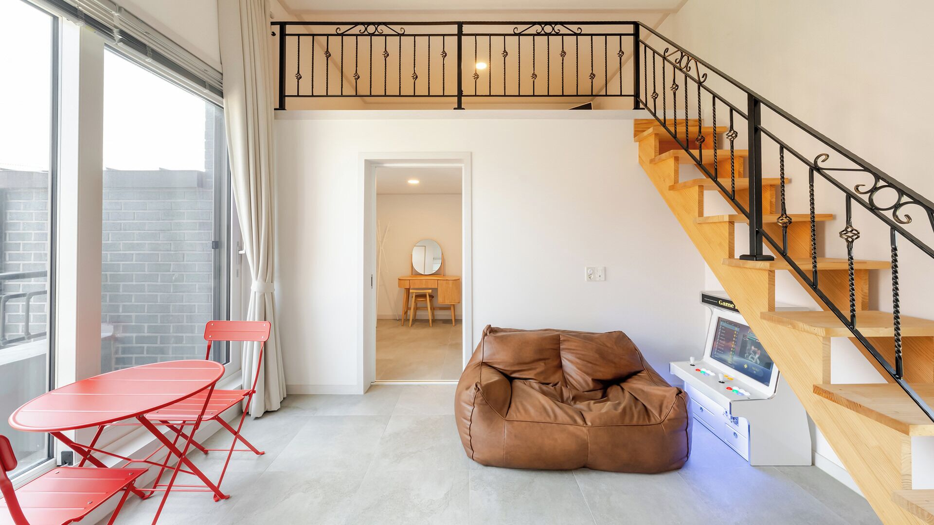 Um apartamento moderno com teto estilo loft, escada de madeira, mesa dobrável vermelha e um puff marrom.