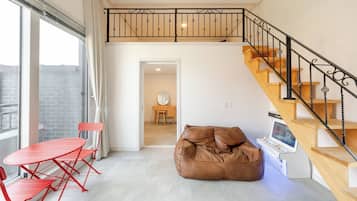 Um apartamento moderno com teto estilo loft, escada de madeira, mesa dobrável vermelha e um puff marrom.