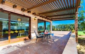 Une terrasse extérieure avec une table et des chaises, un espace salon couvert et une vue sur une piscine et la verdure.