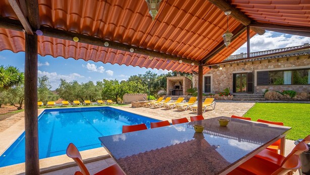 Un área de piscina con un porche cubierto, mobiliario exterior y un edificio de piedra al fondo.