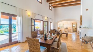 Une salle à manger spacieuse avec un plafond haut et des poutres en bois, de grandes fenêtres offrant une vue sur l’extérieur, et un coin salon confortable avec une cheminée.