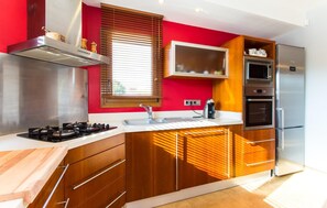 A modern kitchen with a red accent wall, stainless steel appliances, wooden cabinets, and a window with blinds. - Amazing home in Portocolom with private swimming pool, can be inside or outside (Portocolom)