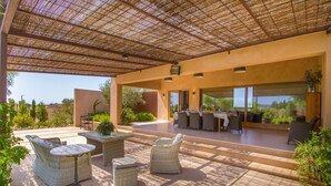 Ein möblierter Patio-Bereich im Freien mit Korbmöbeln, einem überdachten Sitzbereich und Blick auf die umliegende Landschaft.