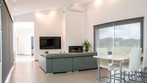 Una sala de estar moderna con un sofá gris, un televisor de pantalla plana, una chimenea y un comedor con sillas blancas y una mesa de vidrio.