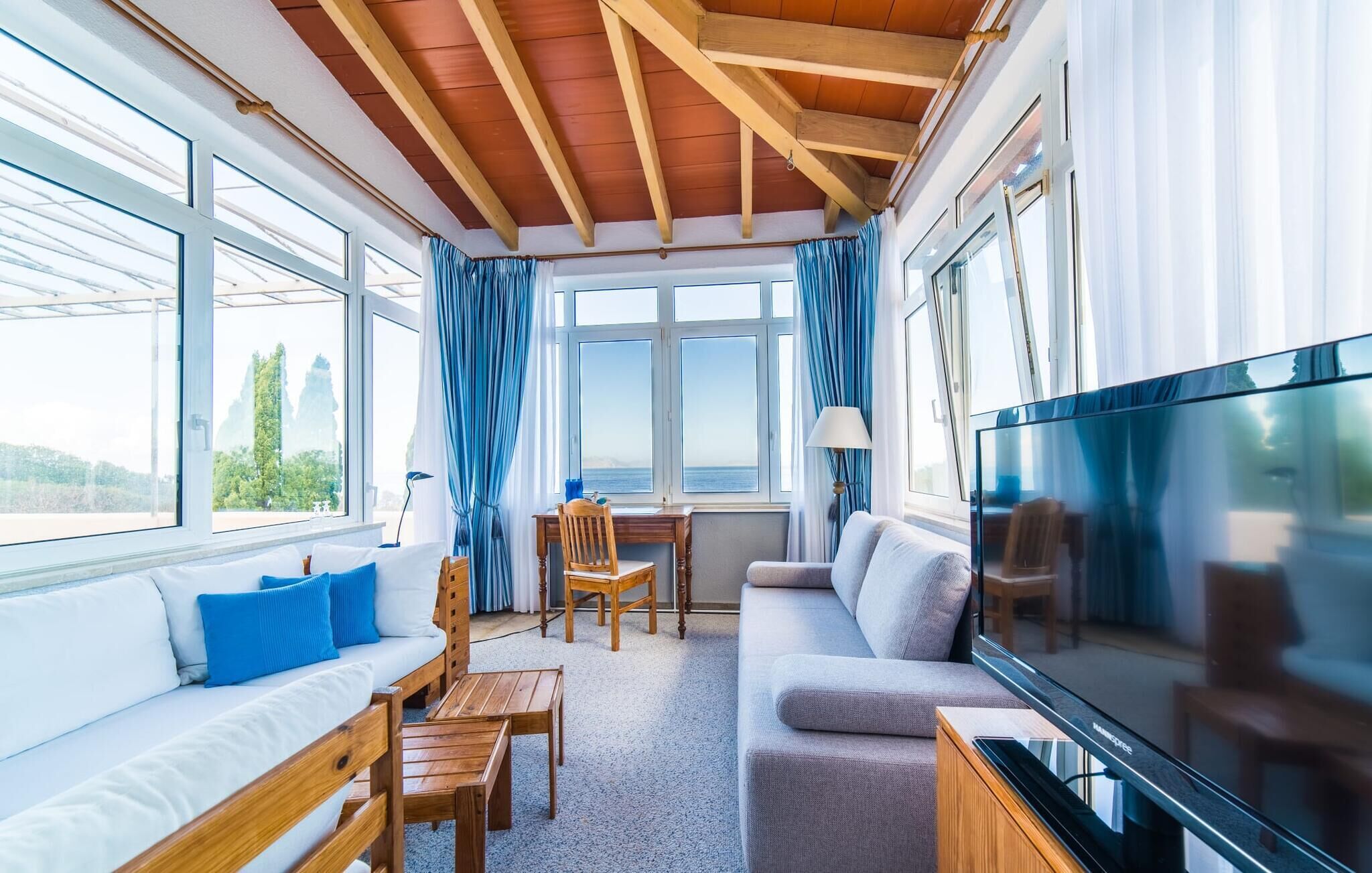 Ein gemütliches Wohnzimmer mit Holzbalkendecke, großen Fenstern, einer Couch, einem Fernseher und einem Essbereich.