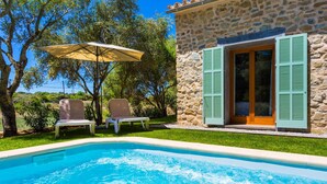 Ein Swimmingpool mit zwei Liegestühlen und einem Sonnenschirm, umgeben von einem Steinhaus mit grünen Fensterläden.