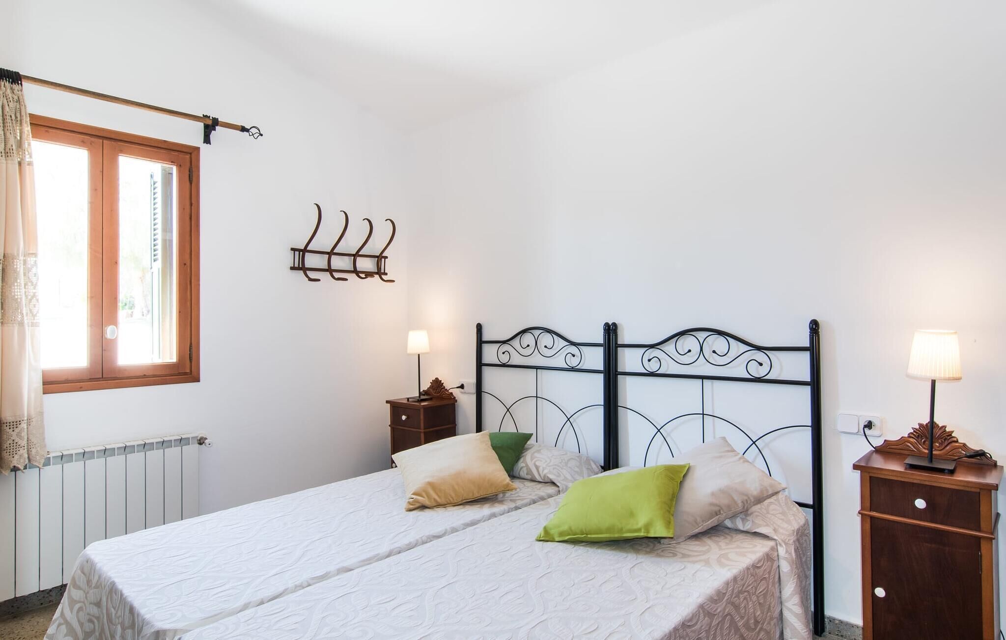 A bedroom with a bed, two bedside tables, a wall-mounted lamp, a window with curtains, and a radiator.