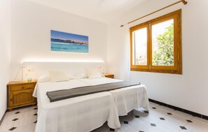Quarto com cama, mesa de cabeceira, janela com vista para vegetação e um quadro de uma praia.