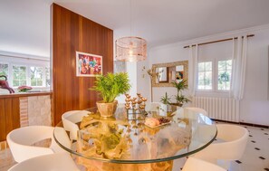 A dining area with a glass table surrounded by white chairs, a wooden accent wall, and a hanging light fixture. - Amazing home in Sa Font de Sa Cala (Sa Font de Sa Cala)